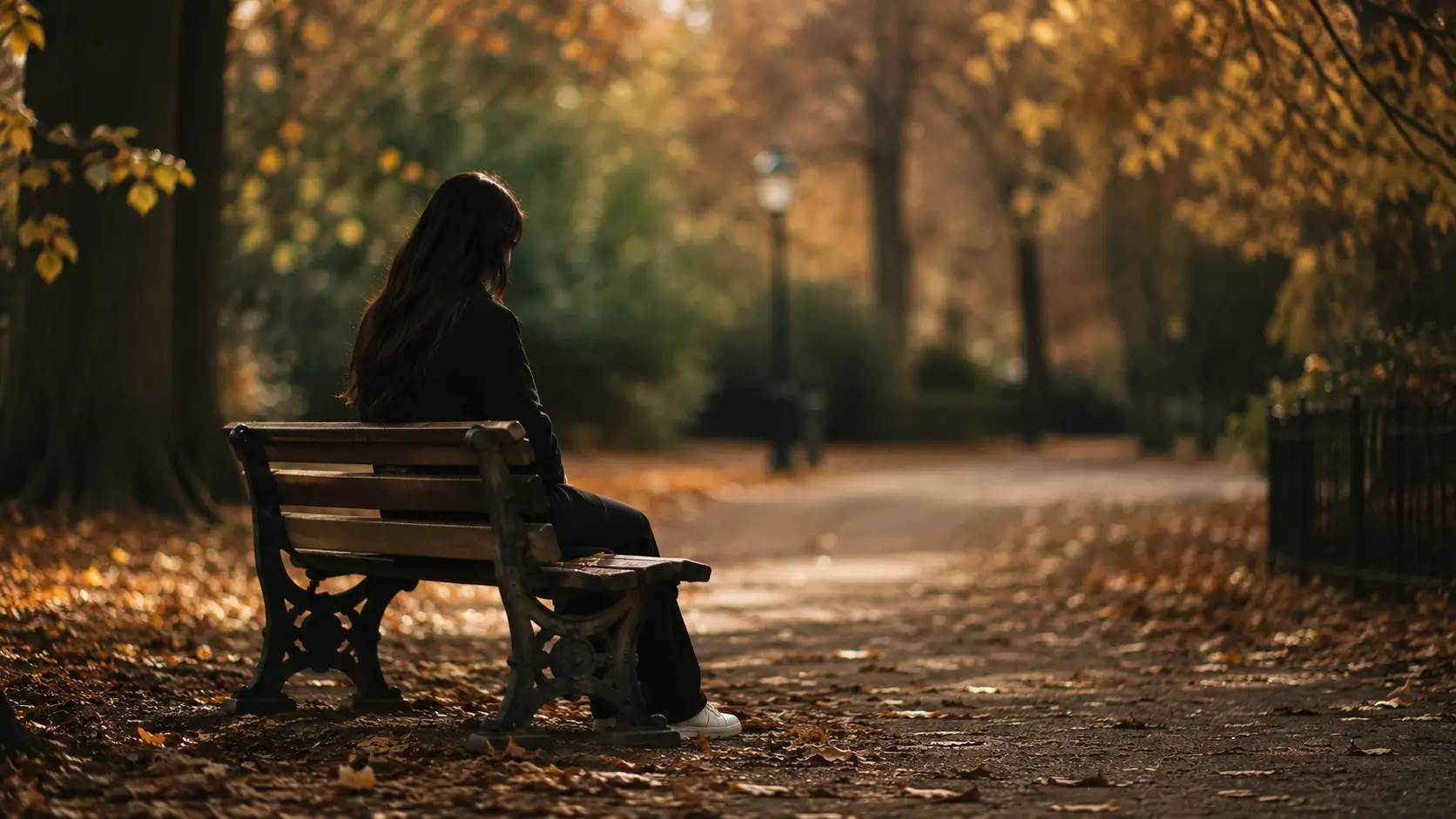 Astrology love cycles — woman sitting alone on park bench in autumn, hands in lap, looking down, fallen leaves around her