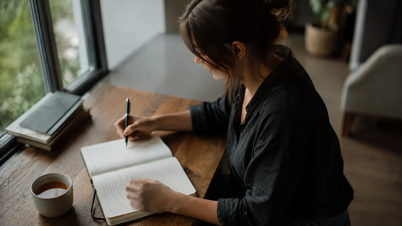 Astrology love testimonials — woman writing in an open journal by a sunlit window, pen in hand, expression calm and focused