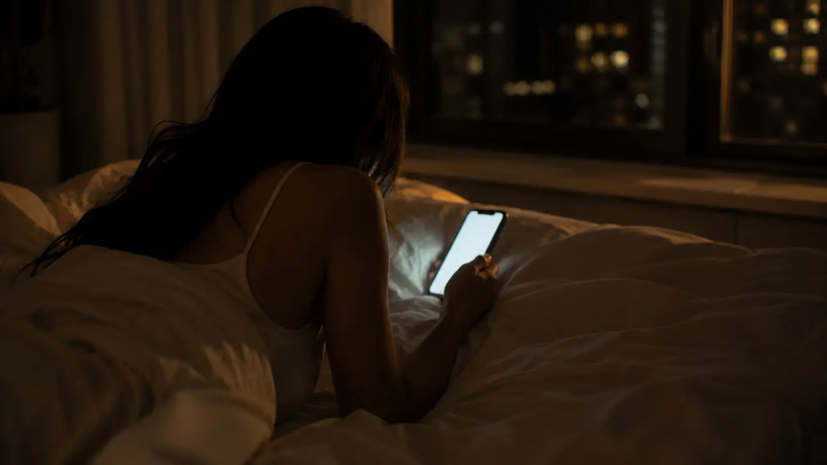 birth chart compatibility love — a woman lying on her side in bed in a dimly lit bedroom, looking at the glow of her phone screen, face not visible, the light soft and quiet around her