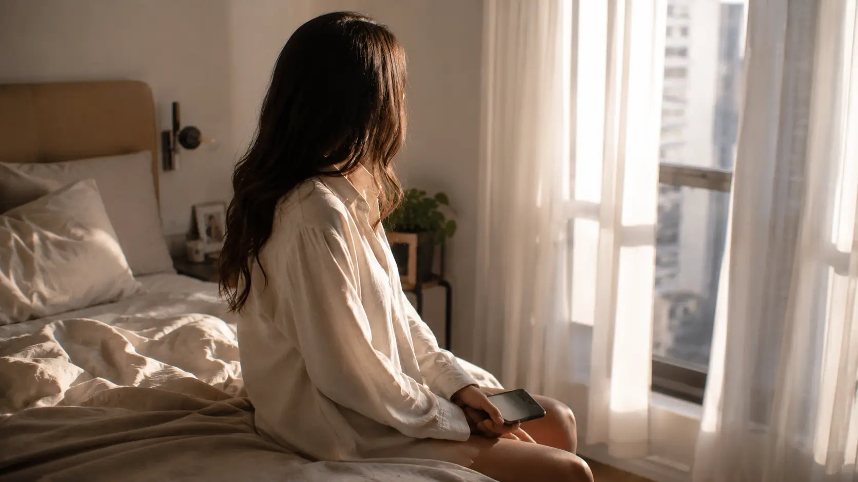 is he the one synastry chart — a woman sitting on the edge of an unmade bed in a softly lit apartment bedroom, holding her phone face-down on her lap, looking toward the window in the early morning light