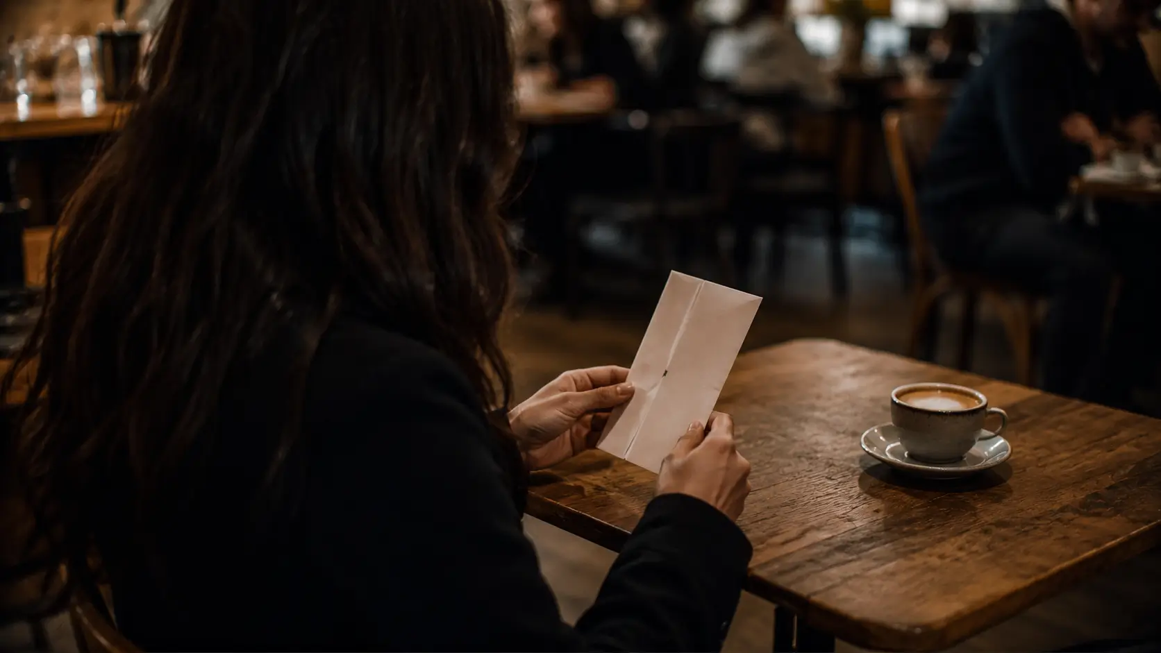 Soulmate drawing astrology reading — woman sitting at a café table holding a folded paper, not yet opened, warm interior light