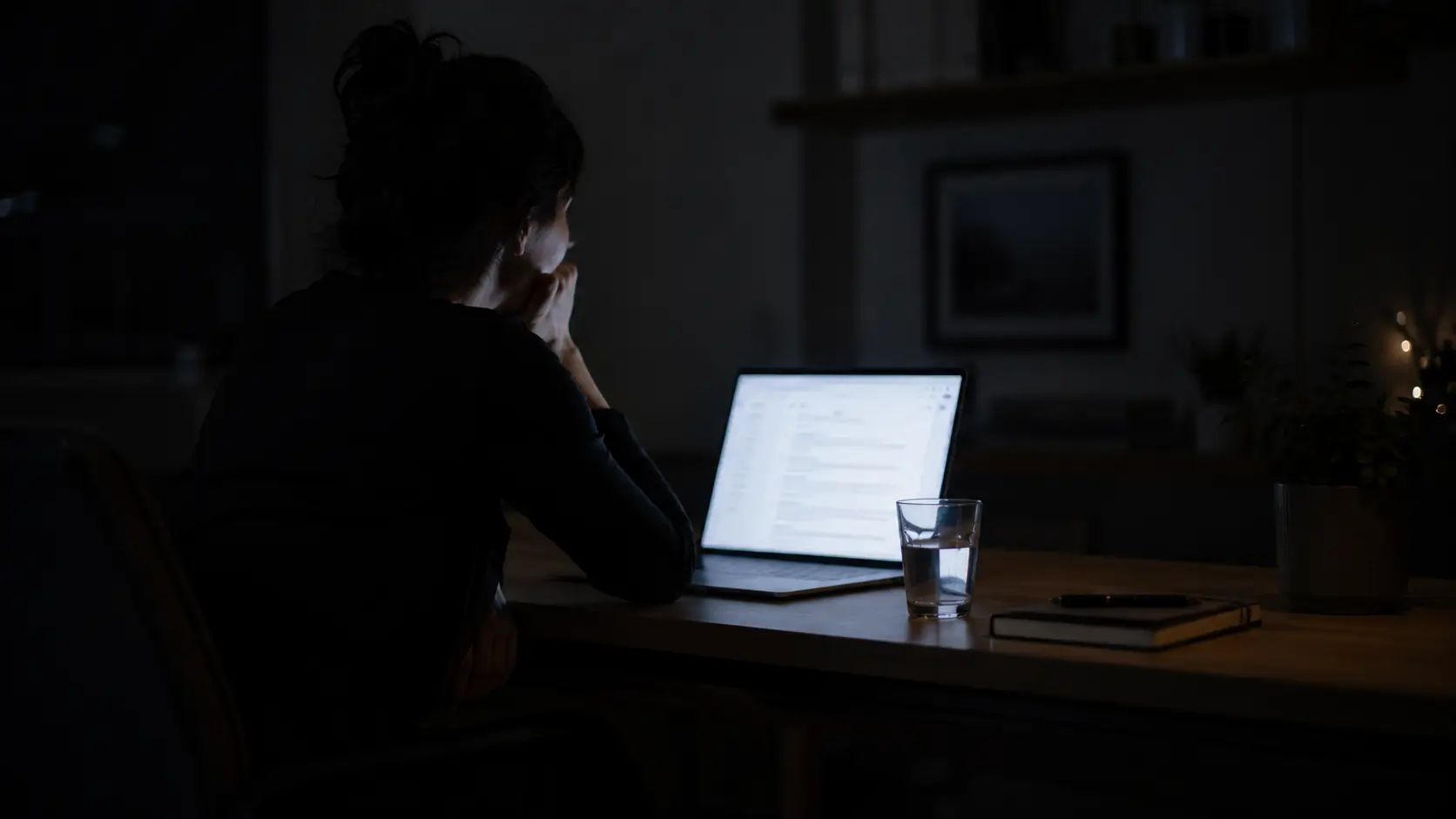 Why i keep repeating the same relationship — woman sitting alone at desk at night, laptop glow, chin resting in hand, not reading