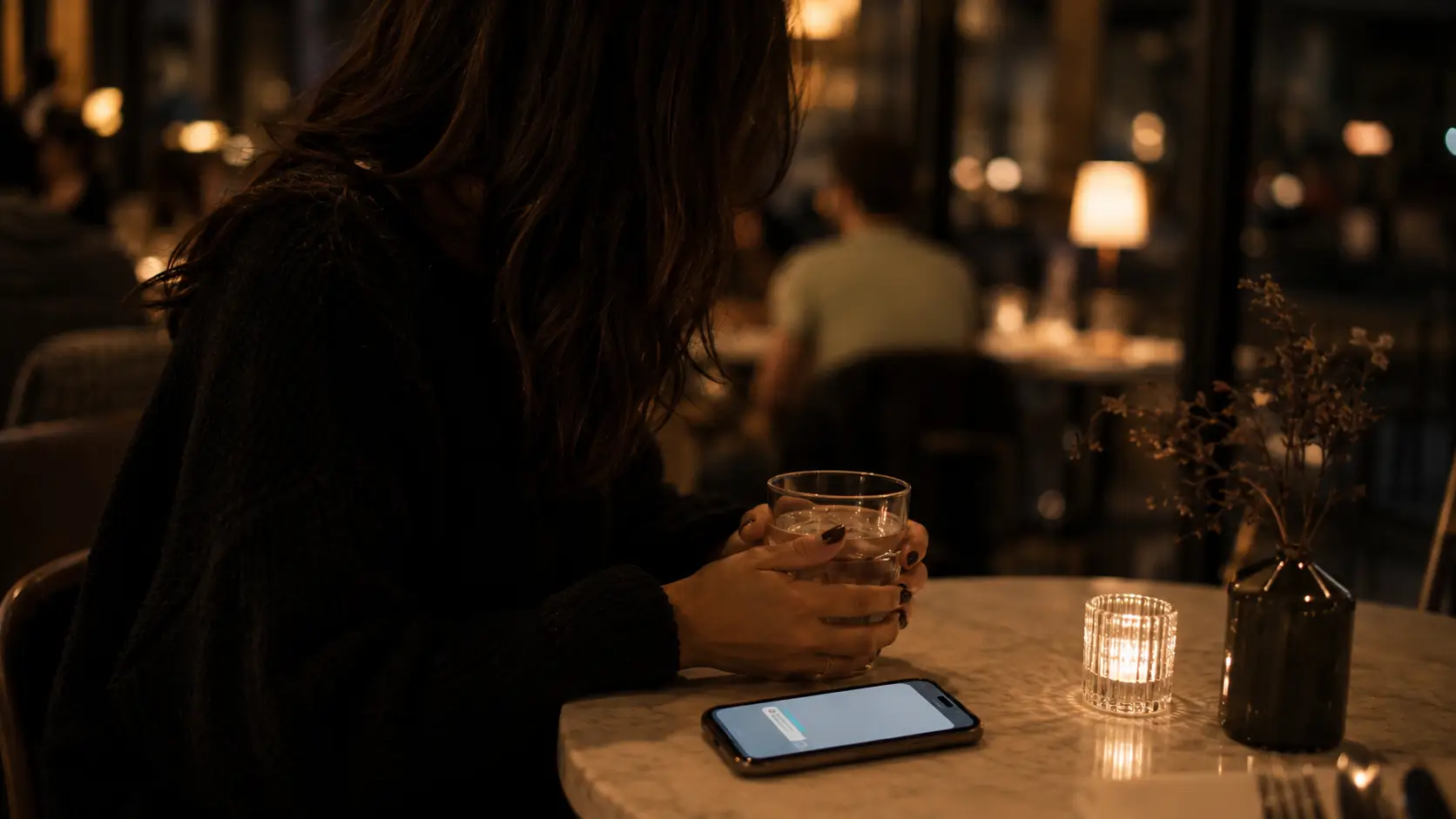 Zodiac sign love patterns — woman sitting at a café at night with a lit phone on the table she hasn’t touched